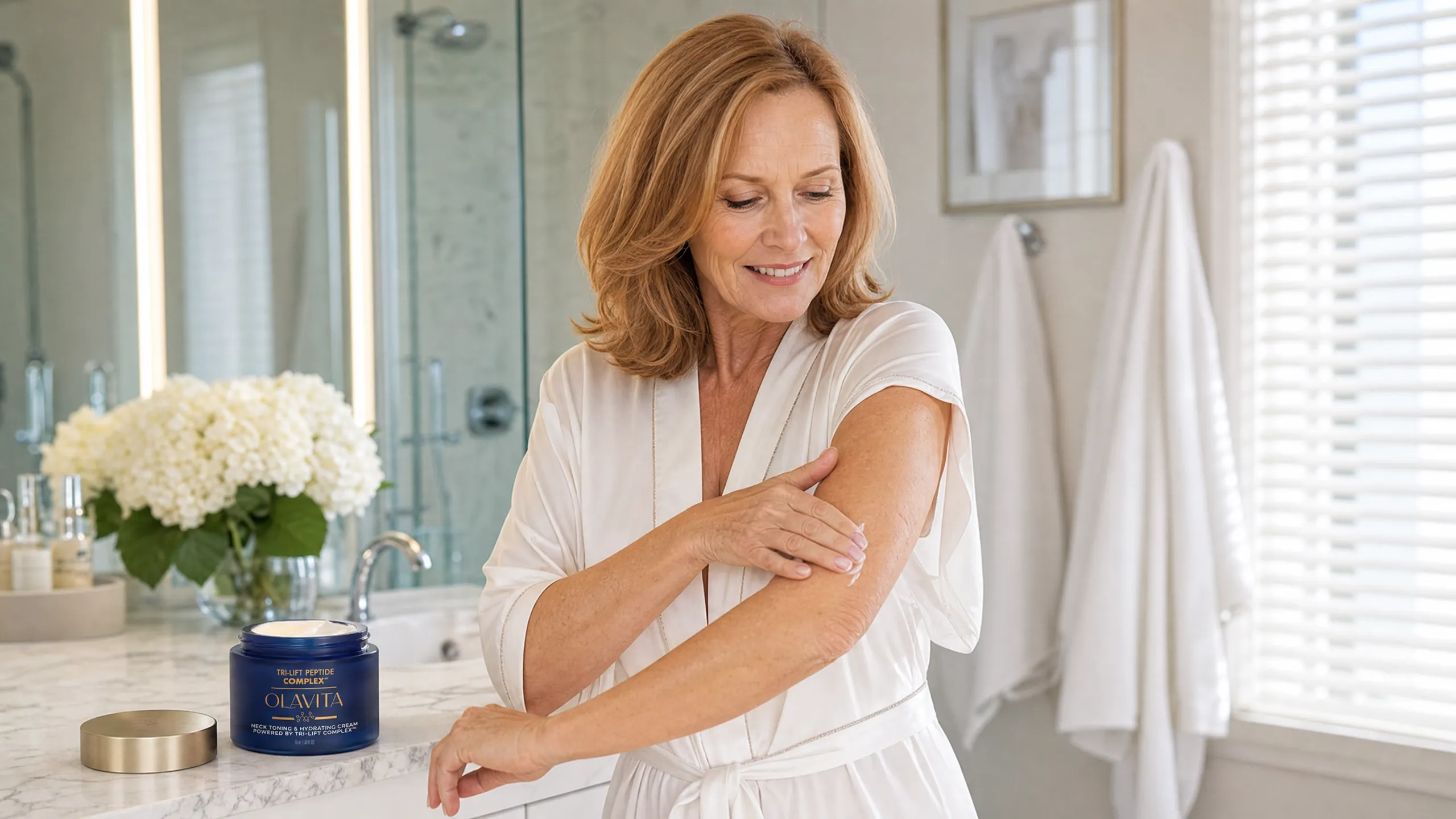A woman in a bathrobe applying cream to her upper arm at a bathroom counter, the jar beside her